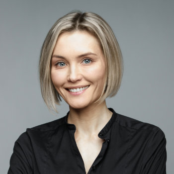 A person with a short blond bob haircut, wearing a black shirt, is smiling against a plain gray background.