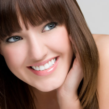 A woman with straight brown hair and bangs smiles brightly, looking to the side with her hand resting near her cheek.
