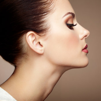 Profile view of a person with a high bun, wearing makeup with defined eyeliner and red lipstick, against a neutral background.