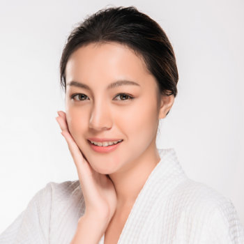 A person with dark hair wearing a white robe, touching their face and smiling against a plain background.