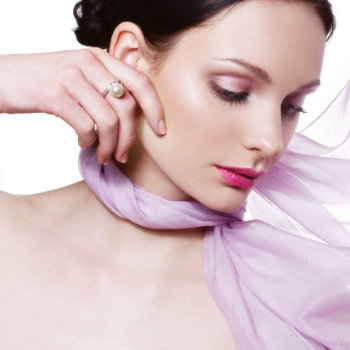 A woman with light skin and dark hair, wearing pink lipstick and eyeshadow, touches her face with one hand. She has a pearl ring and is adorned with a sheer, lavender fabric around her neck.