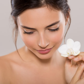 A woman with dark hair holds a white flower close to her face while looking down and slightly smiling.