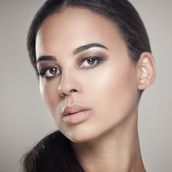 A woman with long dark hair tied back, wearing subtle makeup with smokey eyes and nude lipstick, looks directly at the camera against a plain background.