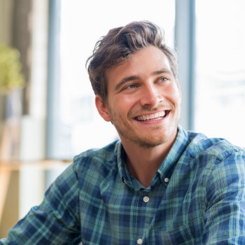A man with short brown hair and a beard, wearing a blue plaid shirt, smiles while seated indoors with windows in the background.