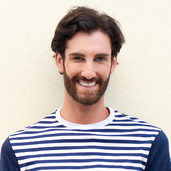 A man with a beard and short hair smiles while wearing a white and navy striped shirt. He stands against a plain, light-colored background.