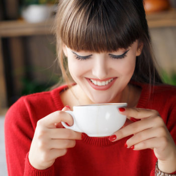 A woman with bangs in a red sweater smiles while holding a white coffee cup near her face, looking down at the cup's contents.