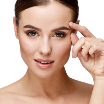 A woman with fair skin and brown hair looks directly into the camera. She is touching her right temple with her index finger. The background is a plain white.