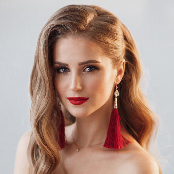 A woman with long, wavy blonde hair, red lipstick, and red tassel earrings looks directly at the camera against a plain background.