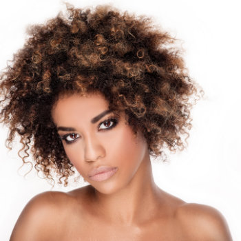 A woman with curly hair styled in an afro poses against a white background. She has natural makeup and a neutral expression.
