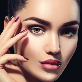 A close-up of a woman with flawless makeup, neatly groomed eyebrows, and glossy lips. She touches her face with fingers manicured in dark polished nails. The background is black.
