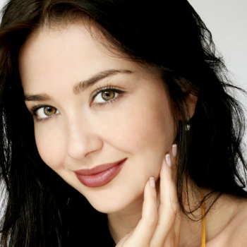 Woman with long dark hair and light skin smiles gently, resting her chin on her hand. She has light eyes and is wearing a yellow top.