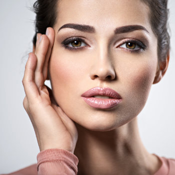 Close-up of a person touching their face with one hand, showing detailed makeup, glossy lips, well-defined eyebrows, and smooth skin. The person is wearing a pink top and looking directly at the camera.