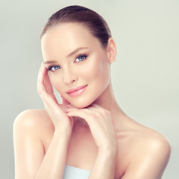 A woman with fair skin and blue eyes lightly touching her face with one hand, smiling, and looking at the camera. She has her hair pulled back and is wearing a strapless white top.