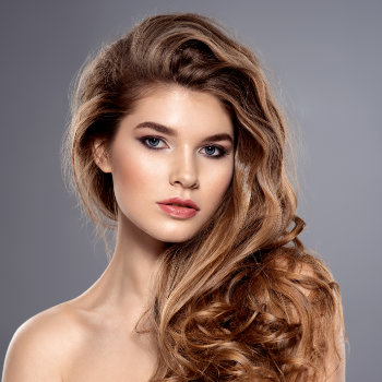 A woman with long, wavy brown hair looks directly at the camera against a gray background. She is wearing natural makeup.