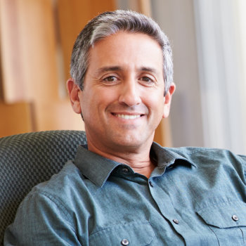 A man with gray hair and a gray shirt smiles while seated in a chair indoors.