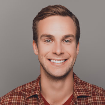 A man with short, brown hair is smiling. He is wearing a red plaid shirt and a red T-shirt underneath, against a plain gray background.
