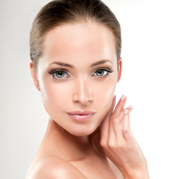 A woman with light skin and brown hair is shown posing with her hand touching her face. She has minimal makeup and is looking directly at the camera against a white background.