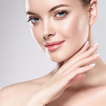 Close-up of a person with fair skin, light blue eyes, and light pink lips, touching their face with manicured fingers. The background is a gradient of light gray.