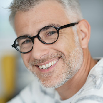 A man with short gray hair, wearing round black glasses and a light-colored shirt, smiles while looking at the camera.