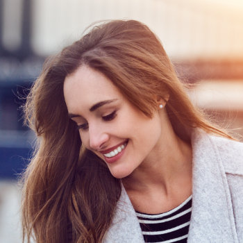A woman with long brown hair, wearing a light gray coat and striped shirt, smiles while looking down.