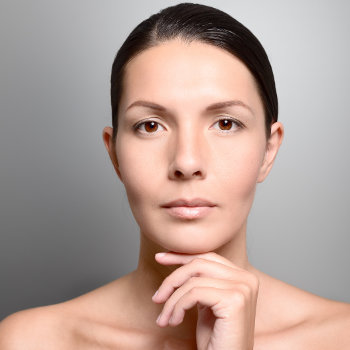 A woman with dark hair pulled back and a neutral expression rests her chin on her hand against a grey background.