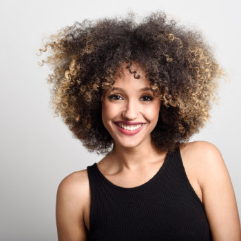 Smiling person with curly hair wearing a black sleeveless top, standing against a plain light gray background.