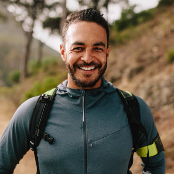 A man wearing a blue jacket and a backpack smiles outdoors with a blurred natural background of trees and hills.