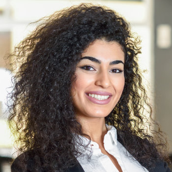A person with curly hair, wearing a white shirt and black blazer, stands smiling in an indoor setting.