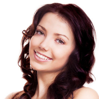 A woman with long curly chestnut hair smiles at the camera. She has light skin and is wearing natural makeup.