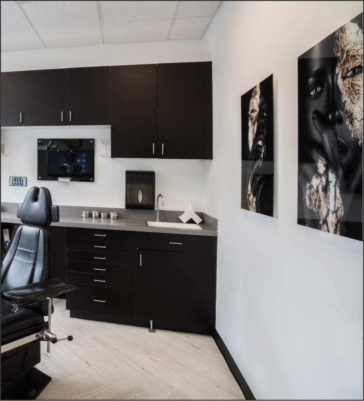 A modern examination room with dark cabinetry, a black medical chair, a countertop sink, wall-mounted TV, and two artistic portraits on the wall. The room has a clean and minimalist design.