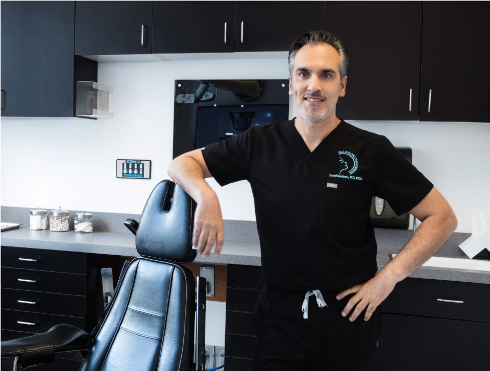 A person wearing black scrubs stands next to a chair in a dental office, with cabinets and equipment in the background.