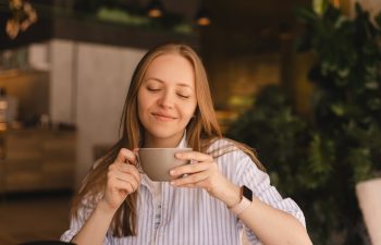 Portrait of gorgeous blonde smiling lady smelling, enjoying of coffee and drinking cappuccino from cup while resting in restaurant. Woman wear stripped shirt.